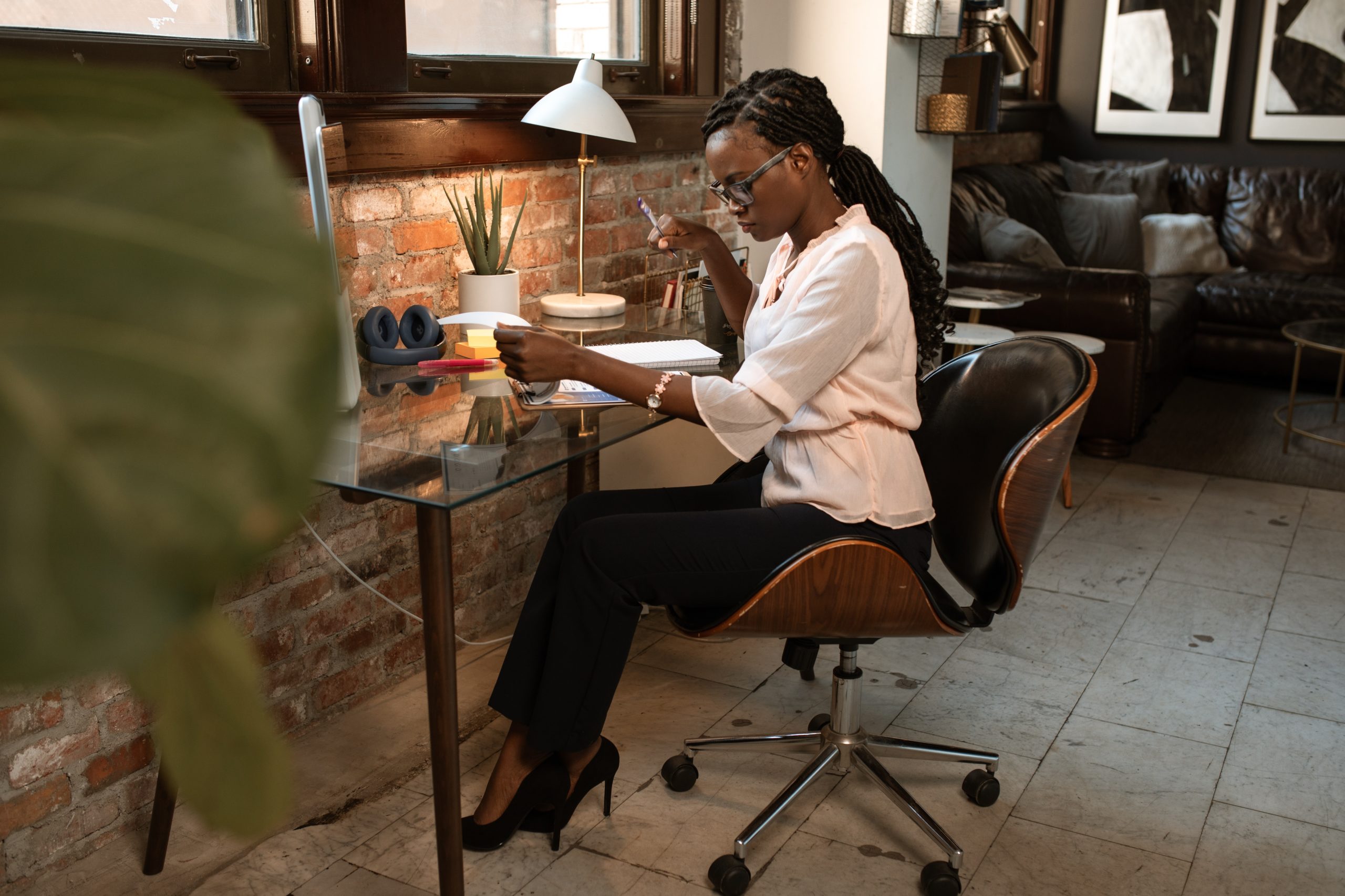 A person with long braids, glasses, and high heels sits at a desk in a modern office space, reading documents and taking notes. The desk has a computer, lamp, and other office supplies. The background includes a brick wall, large window, and a couch.