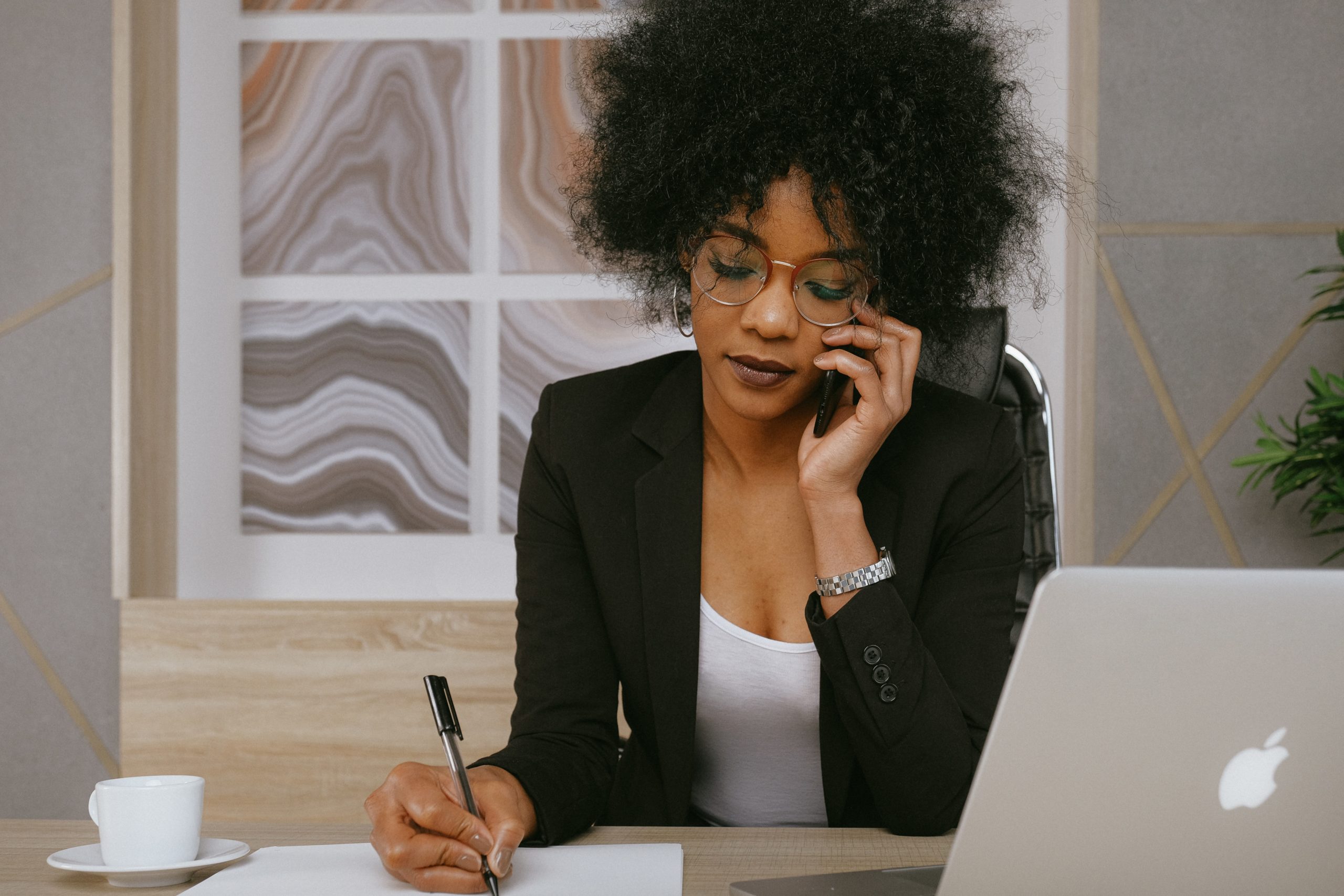 A woman with curly hair and glasses is sitting at a desk, talking on her phone while writing on a piece of paper. She is dressed in a black blazer and white blouse. A cup of coffee, a laptop, and a plant are on the desk. Abstract artwork is in the background.