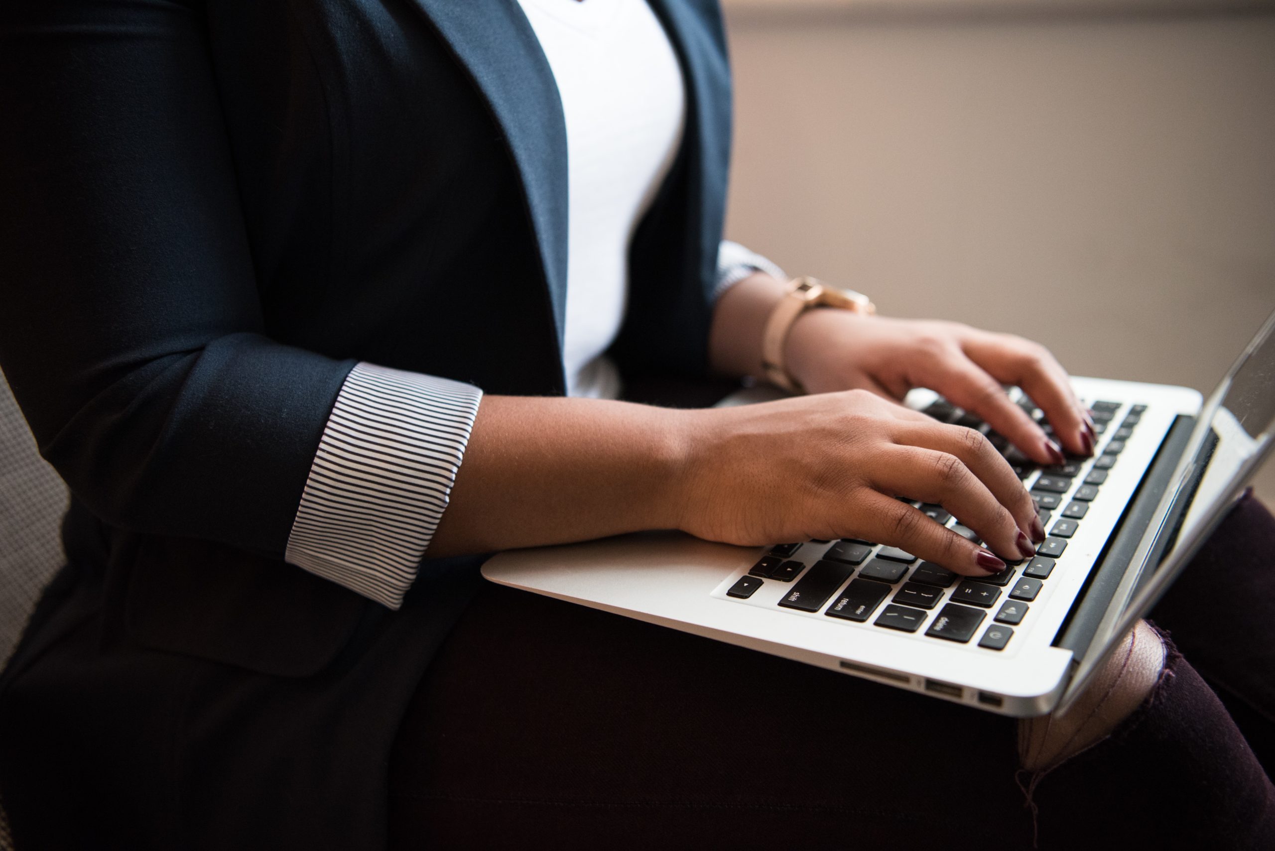 A person wearing a black blazer and a white shirt types on a laptop. Their hands rest on the keyboard, with focus on their fingers. The background is blurred, emphasizing the laptop and the person's hands.
