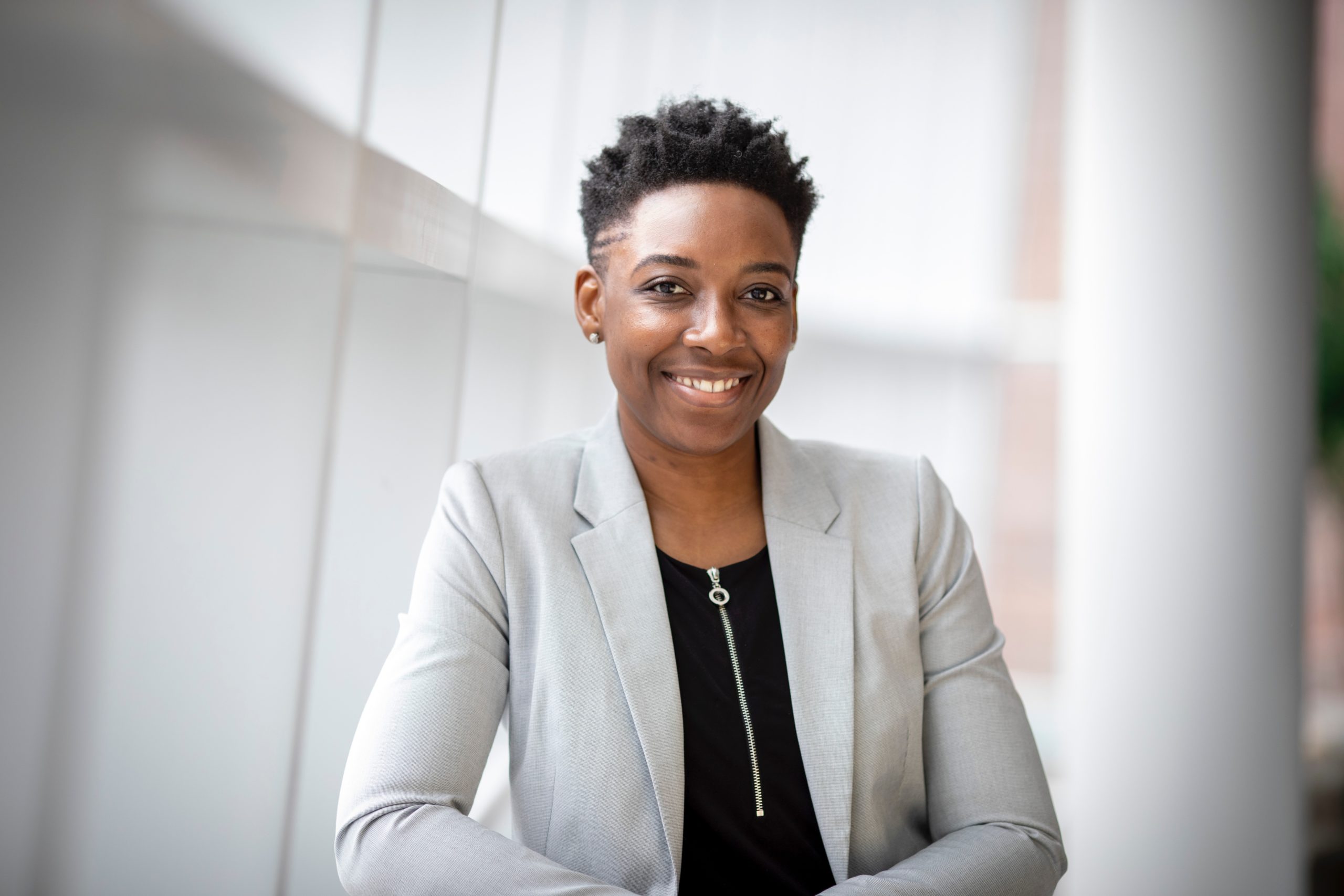 A person with short hair and a friendly smile is wearing a light gray blazer over a black shirt. The background is a bright, softly lit indoor space with white walls and windows.