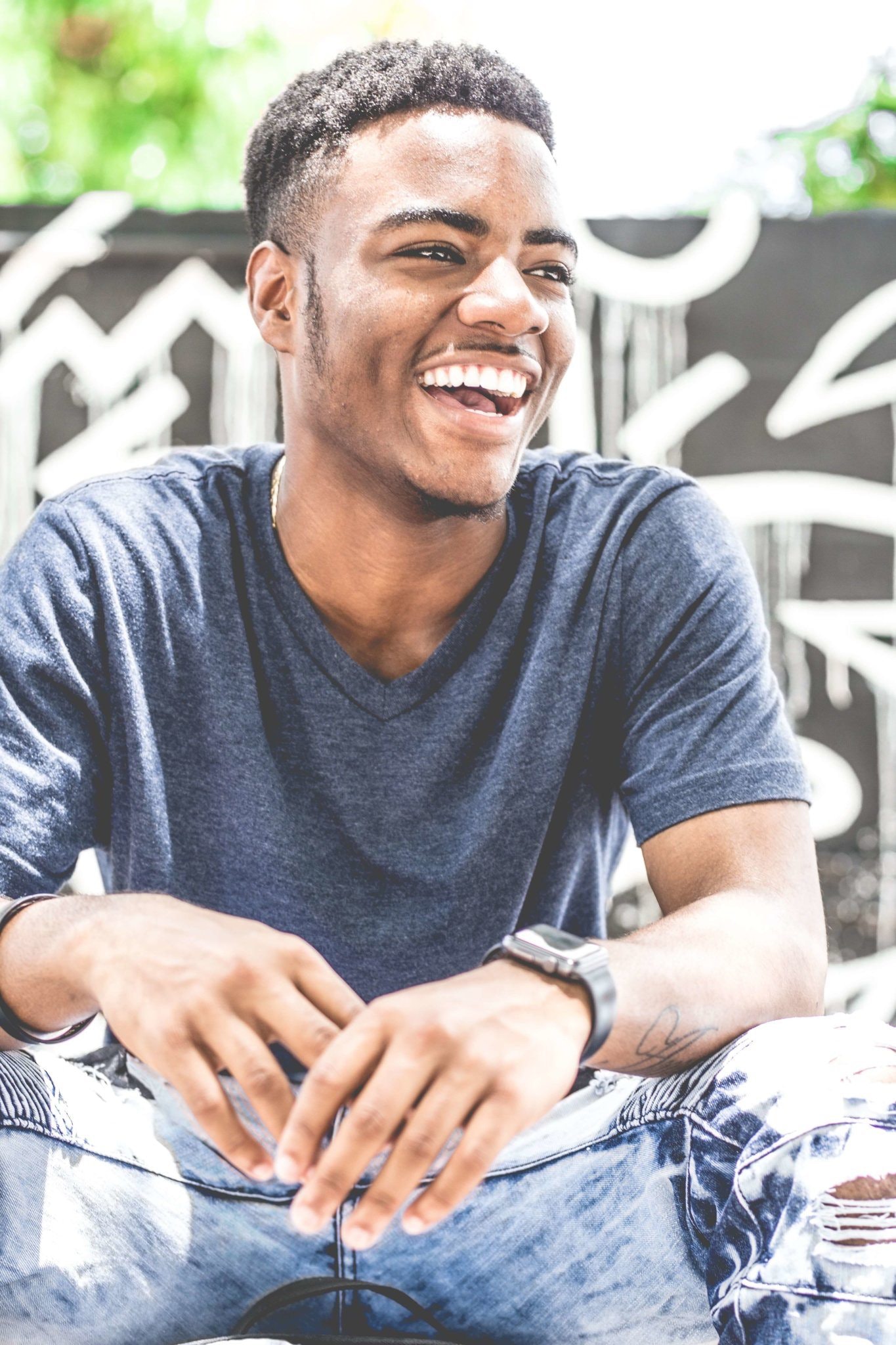 A young man with short curly hair is sitting outdoors and laughing. He is wearing a dark gray V-neck T-shirt and light blue jeans, with a modern watch on his left wrist. The background has an artistic graffiti wall with abstract black and white designs.