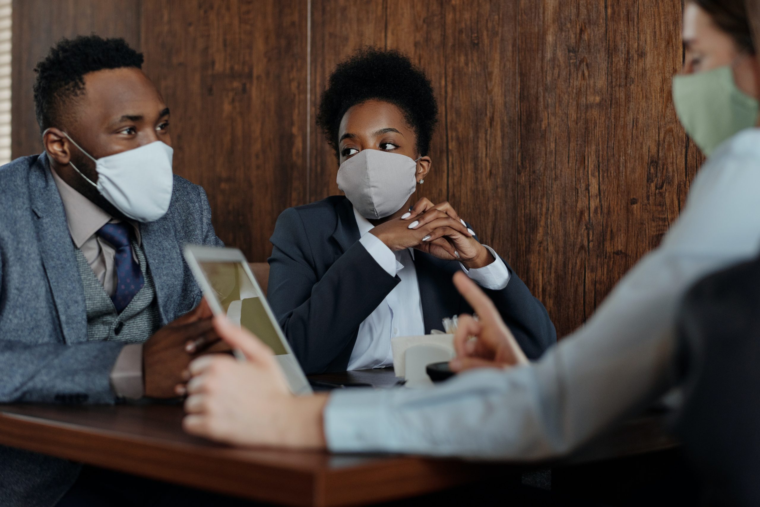 Three people in business attire, wearing face masks, are sitting at a table in a meeting. One person is holding a tablet, and everyone appears engaged in conversation. The background features a wooden wall.