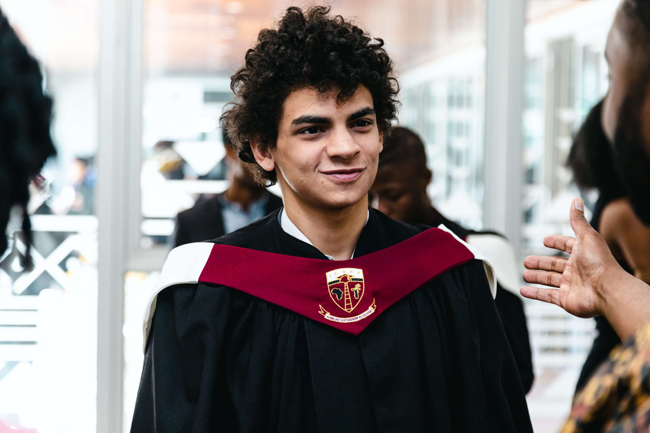 A young person with curly hair smiles while wearing a graduation gown with a red and white sash. They are engaged in conversation with someone whose hand is gesturing in the foreground. The background shows other people and a bright indoor setting. Aamen Muharram: My Time at ALA has Unleashed My Passions.