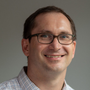 A man with short brown hair and glasses smiles at the camera. He is wearing a checkered shirt and the background is plain and gray.