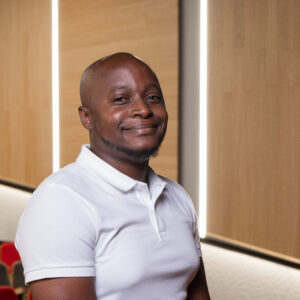 A person with a shaved head smiles warmly while posing for a photo. They are wearing a white polo shirt and standing in front of a modern, warmly lit interior with wood paneling and red seating in the background.