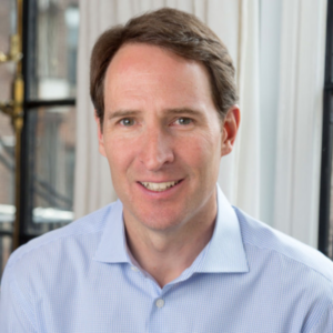 A smiling man with short brown hair is wearing a light blue dress shirt. He is seated in front of a window with white curtains and a black window frame. The background shows an out-of-focus urban setting.
