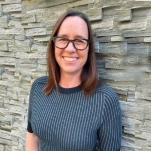 A woman with straight, shoulder-length hair and black-rimmed glasses is smiling while standing against a textured stone wall. She is wearing a dark, short-sleeve sweater with horizontal stripes.