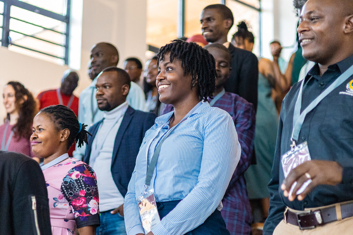 A diverse group of people stands together in a well-lit indoor space. They are smiling and appear to be engaged in an event. Some individuals are wearing name badges and lanyards. The background shows more people and large windows.