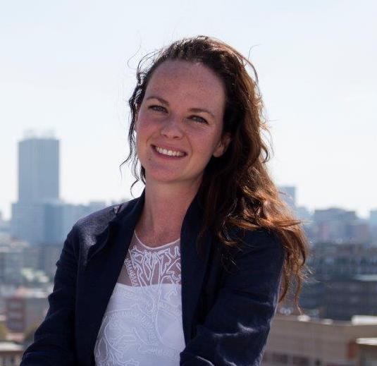A woman with long, wavy hair stands outdoors with a cityscape in the background. She is wearing a dark blazer over a white, patterned top and is smiling at the camera. The sky is clear, and buildings are visible in the distance.