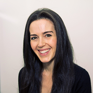 A woman with long dark hair and a friendly smile is posing against a plain, light-colored background. She is wearing a dark top and appears to be looking into the camera.