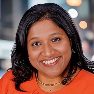 A smiling woman with long dark hair is wearing an orange top and a necklace. She is seen indoors with a blurred background of lights and people.