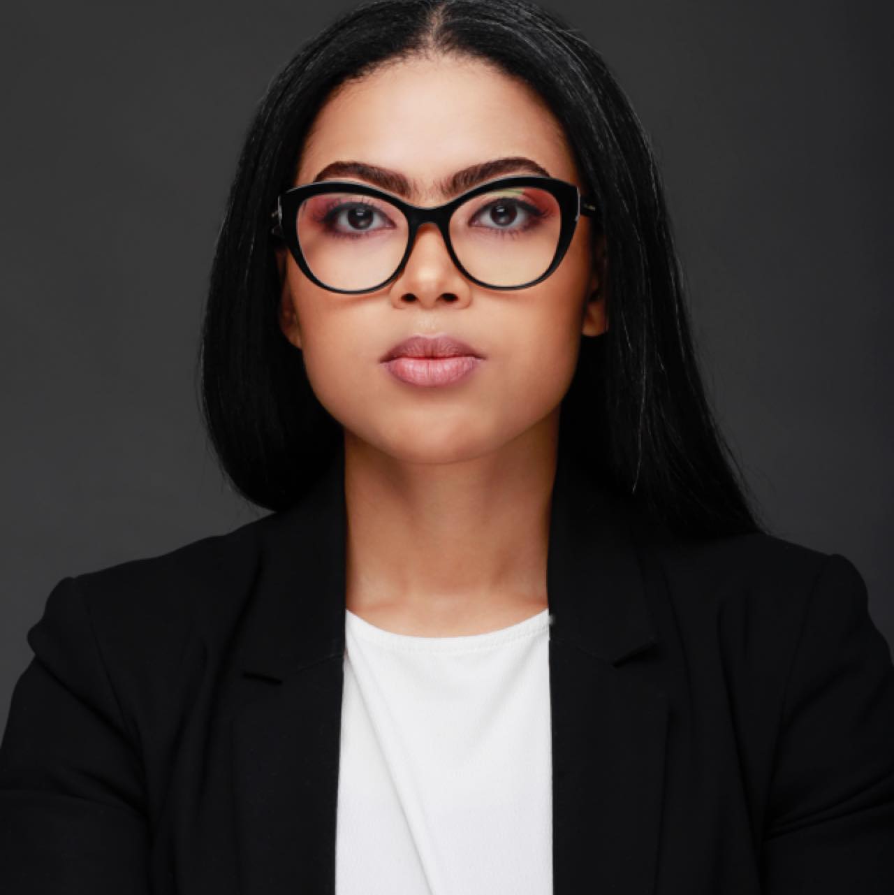 A woman with long dark hair wearing oversized black glasses, a white shirt, and a black blazer is looking directly at the camera with a serious expression. The background is a solid dark gray.