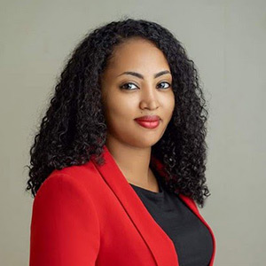 A woman with long curly hair is wearing a red blazer over a black top. She is standing against a plain beige background, looking confidently at the camera with a slight smile.