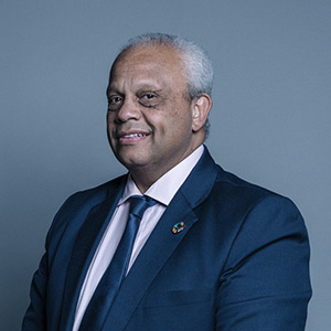 A man with short gray hair, light skin, and a slight smile is wearing a navy suit jacket, white dress shirt, and dark tie. He stands against a gray background. A colorful circular pin is on his jacket lapel.