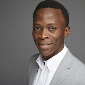 A man in a light gray suit jacket and white dress shirt is smiling slightly, standing against a plain gray background. He has short black hair and appears to be in a professional setting.