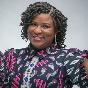 A woman with curly hair smiles warmly at the camera. She is wearing a patterned blouse with geometric designs in pink and blue. Her earrings are visible, and the background is a simple light gray.