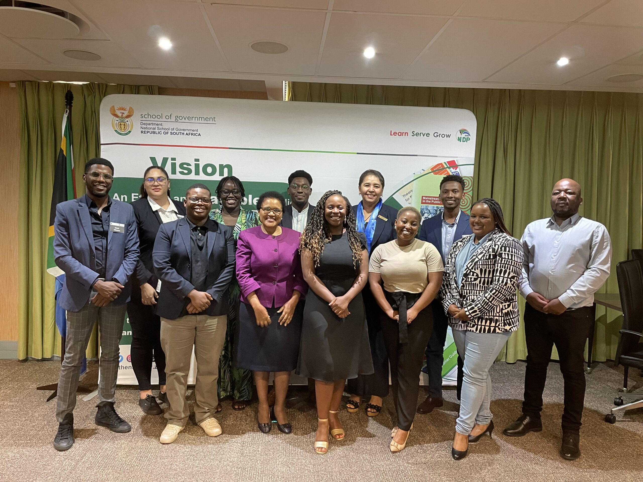 A group of thirteen people posing for a photo in front of a banner that says "Vision: An ethical and capable state" with logos and a government symbol. They are dressed in business attire, standing in a conference room with a South African flag visible.