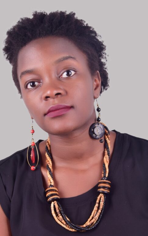 A person with short curly hair wears a black top, a multistrand necklace with black and beige beads, and mismatched earrings featuring a red bead and a black disc. They have a neutral facial expression and are looking slightly upwards. The background is plain gray.