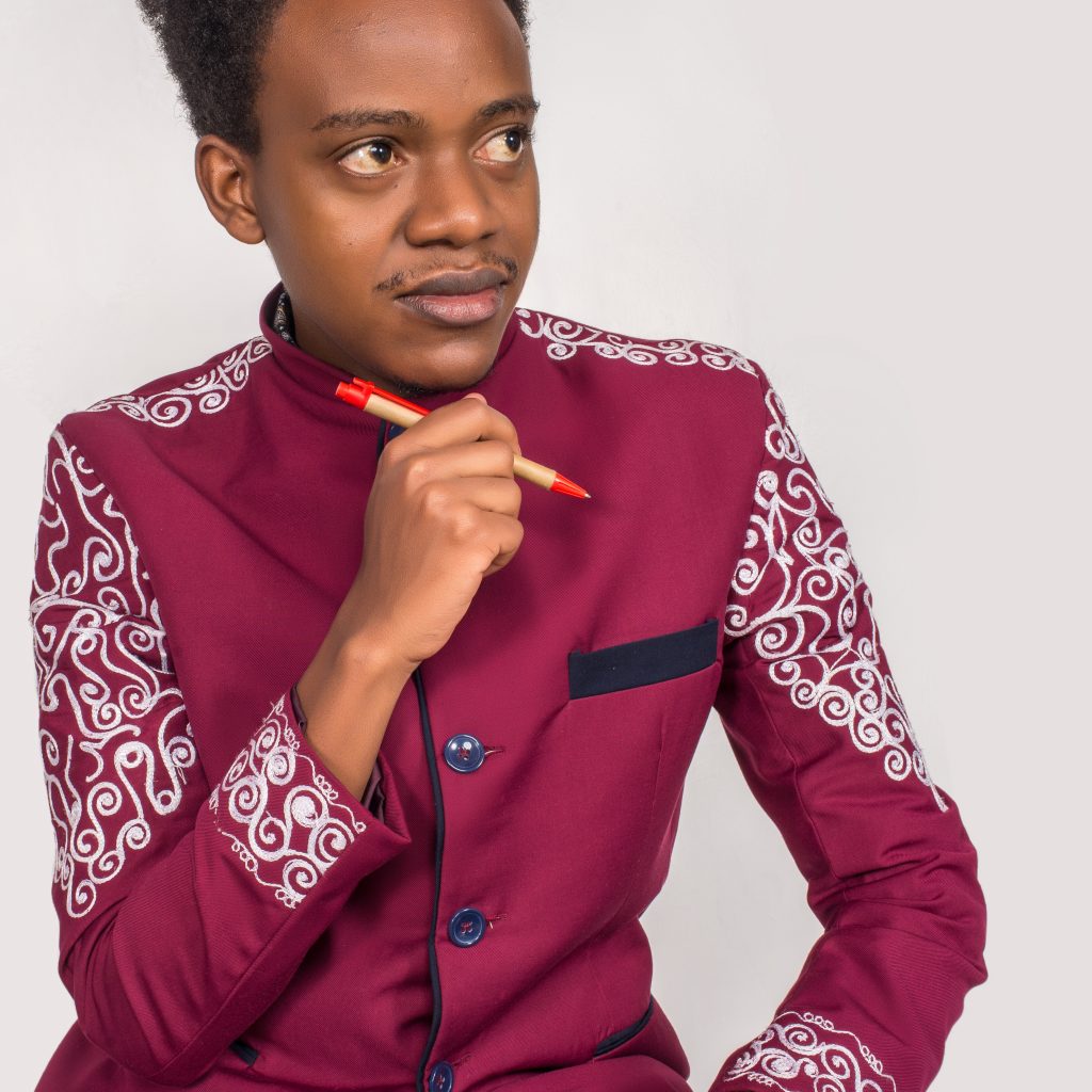 A person wearing a maroon jacket with intricate white patterns on the shoulders and sleeves is holding a red pen near their chin, looking thoughtfully to the side against a plain background.