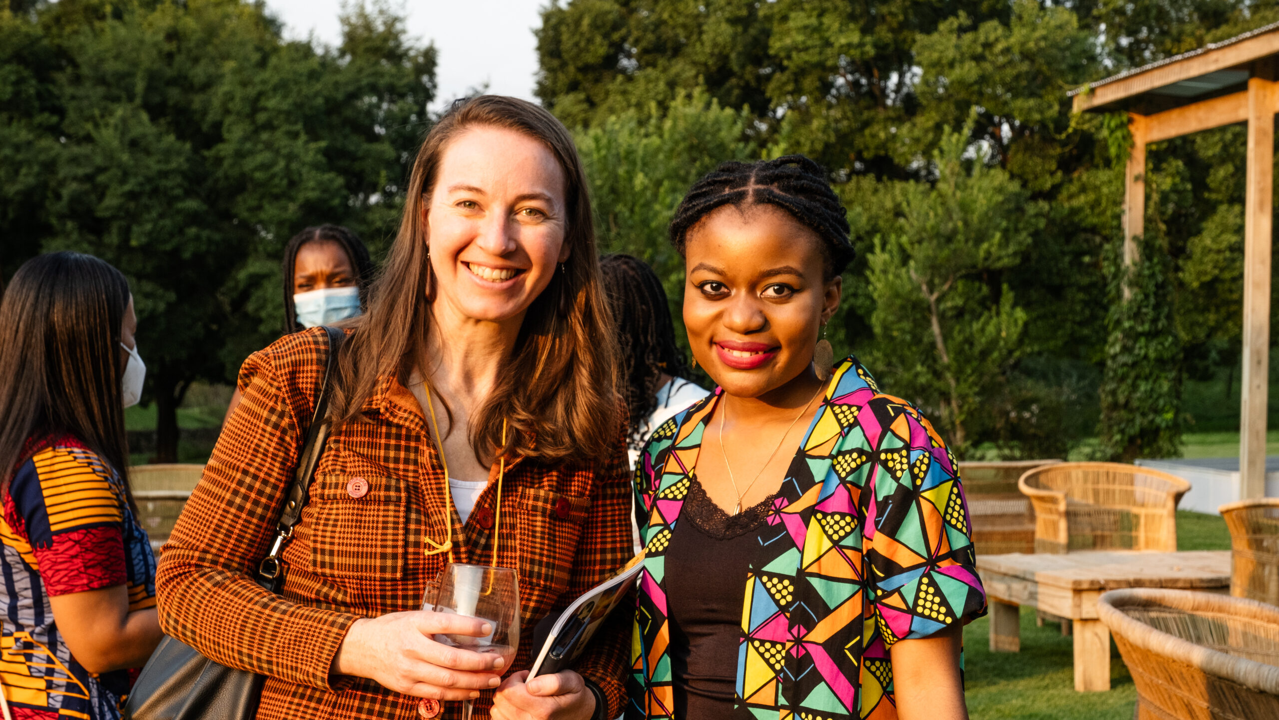Two women are standing outdoors, smiling at the camera. One woman is holding a drink and wearing a brown checked shirt, and the other woman is wearing a colorful, patterned top. People and seating areas are visible in the background, surrounded by greenery.