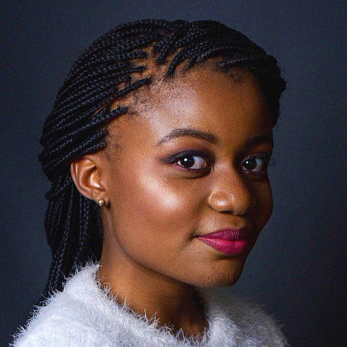 A woman with braided hair, wearing a white fuzzy sweater, smiles softly against a dark background. She has natural makeup with pink lipstick and stud earrings.