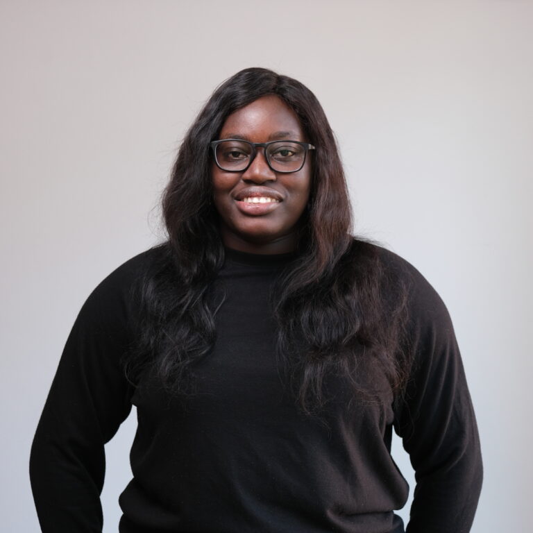 A person with long, dark hair and glasses, smiling at the camera. They are wearing a black long-sleeve shirt and stand in front of a plain, light-colored background.