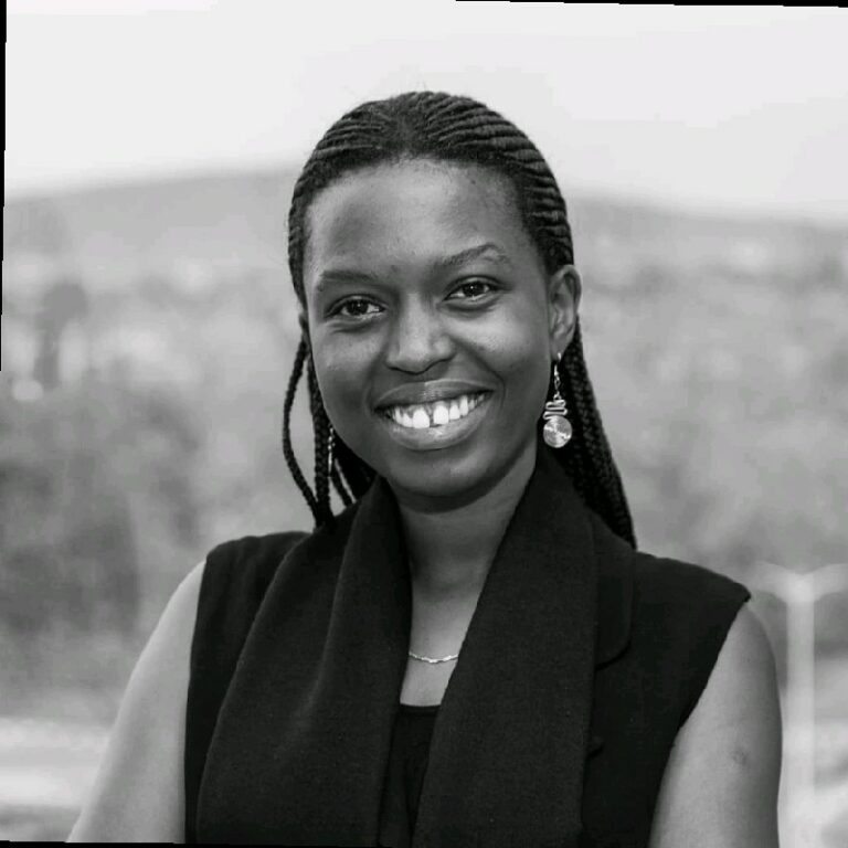 Black and white photo of a person with braided hair smiling at the camera. They are wearing a black sleeveless top and an earring. The background appears to be outdoors with a blurred landscape.