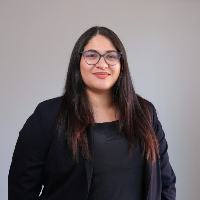 A woman with long dark hair and glasses stands in front of a plain background. She is wearing a black blazer over a black top and is smiling at the camera.