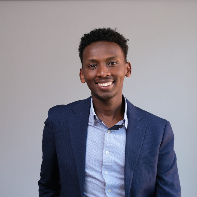 A person stands against a plain backdrop, smiling and wearing a dark blue blazer over a light blue button-up shirt. They have short, curly hair and an overall cheerful expression.