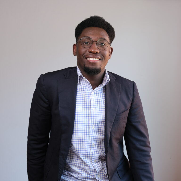 A person with short, curly hair, wearing glasses, a dark blazer, and a checkered shirt smiles while posing against a plain light gray background.