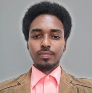 A person with short curly black hair and a beard is seen wearing a light pink shirt and a tan jacket. They are facing the camera with a neutral expression. The background is gray.