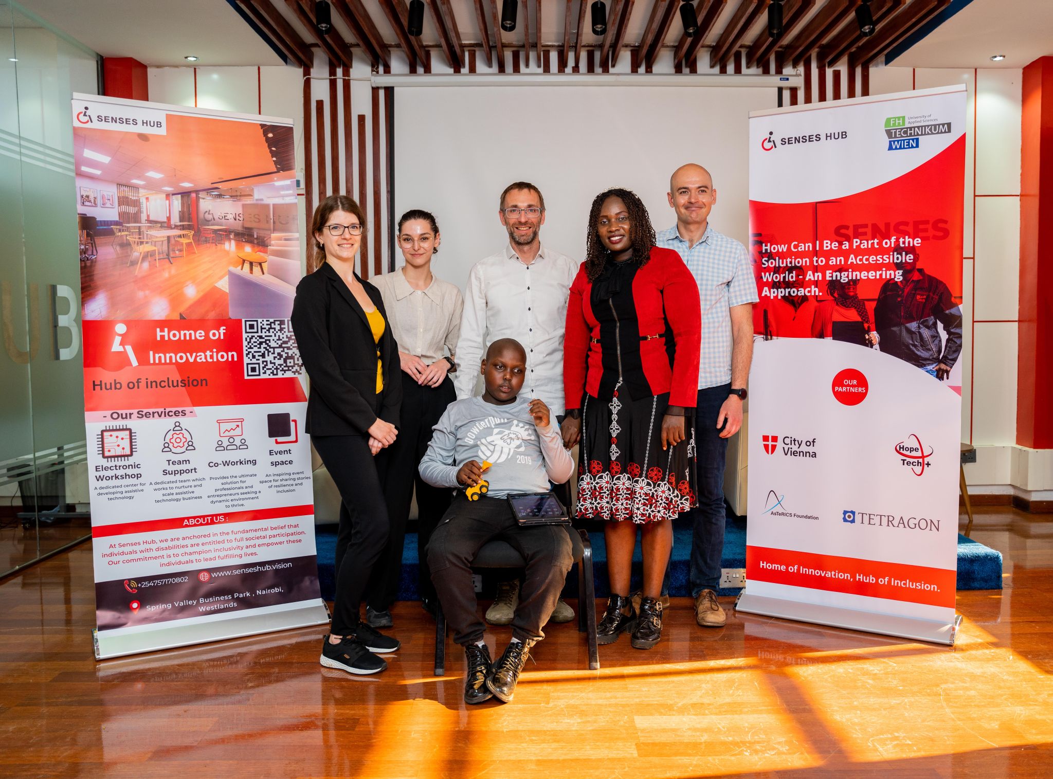 A group of six people posing indoors. Two pull-up banners with text and images are on each side of the group. A person in a wheelchair is seated in the front center, while the other five, dressed in business casual attire, stand around them smiling.