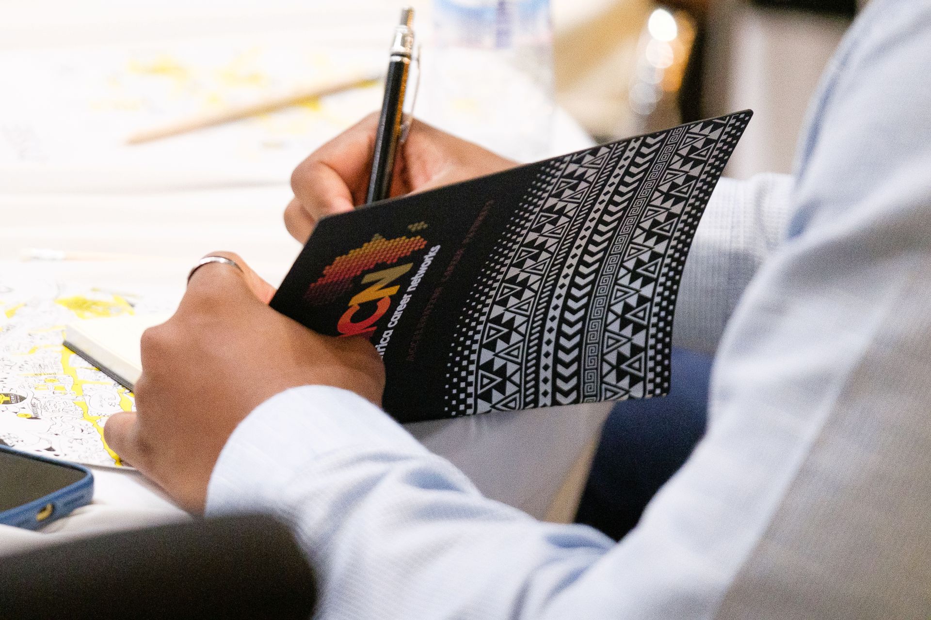 A person in a light blue shirt writes in a black notebook decorated with geometric patterns and a colorful logo, sitting at a desk with papers and a phone nearby.