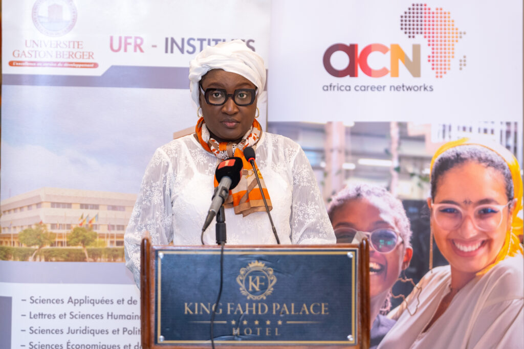 A person in a white outfit and headscarf speaks at a podium labeled "King Fahd Palace Hotel." Behind them are banners for "Université Gaston Berger" and "Africa Career Networks." The setting seems to be a formal event or conference.