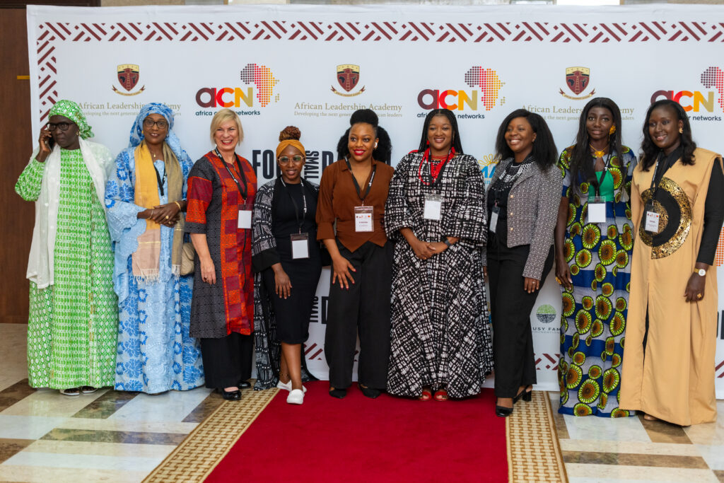 A diverse group of ten people stand on a red carpet in front of a backdrop with logos of African Leaders Academy and ACN. They are smiling and dressed in a variety of colorful, formal, and traditional outfits.
