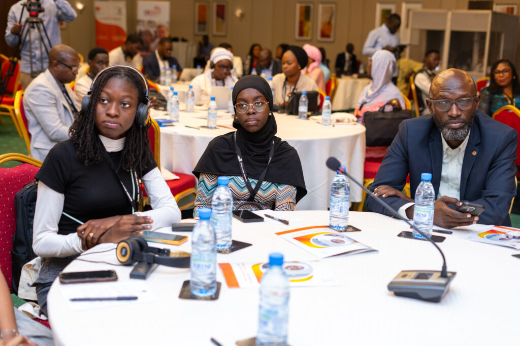 A group of people, including two women and a man, sit attentively at a round table during a conference. They have documents and water bottles in front of them. Other attendees and tables are visible in the background.