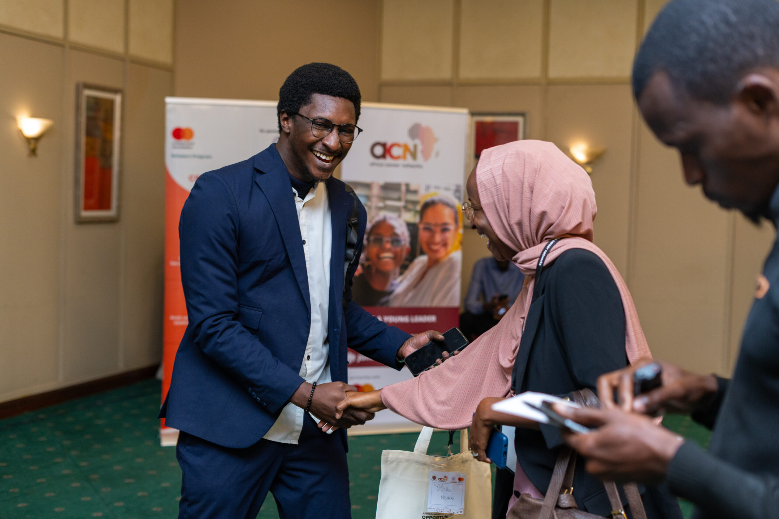 A man in a suit and a woman in a hijab shake hands while smiling at an event. There are people in the background, and a banner with logos. The setting appears to be a conference room.
