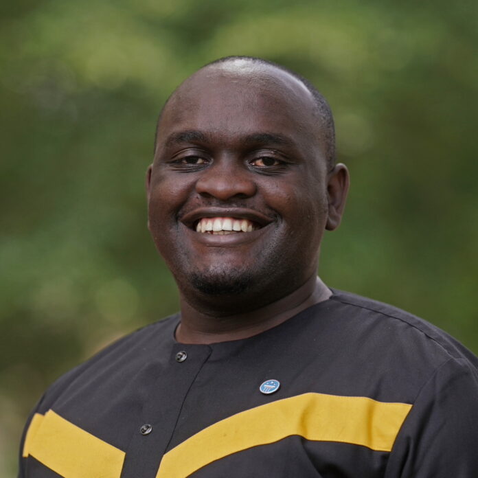 A person is smiling while wearing a black shirt with yellow stripes and a small pin. The background is a blurred green, suggesting an outdoor setting.