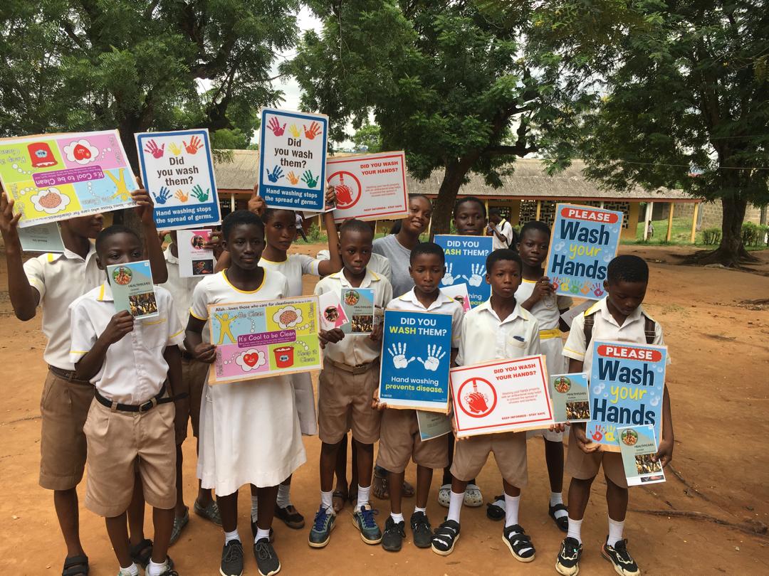 A group of children in school uniforms stand outdoors holding colorful posters about handwashing. The posters feature messages like "Wash your hands" and various illustrations. Trees and a thatched-roof building are in the background.