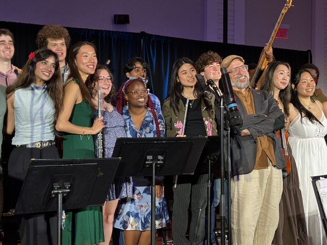 A diverse group of people, some holding musical instruments, stand close together on stage smiling for a photo. Music stands are in front of them and a black curtain is in the background.
