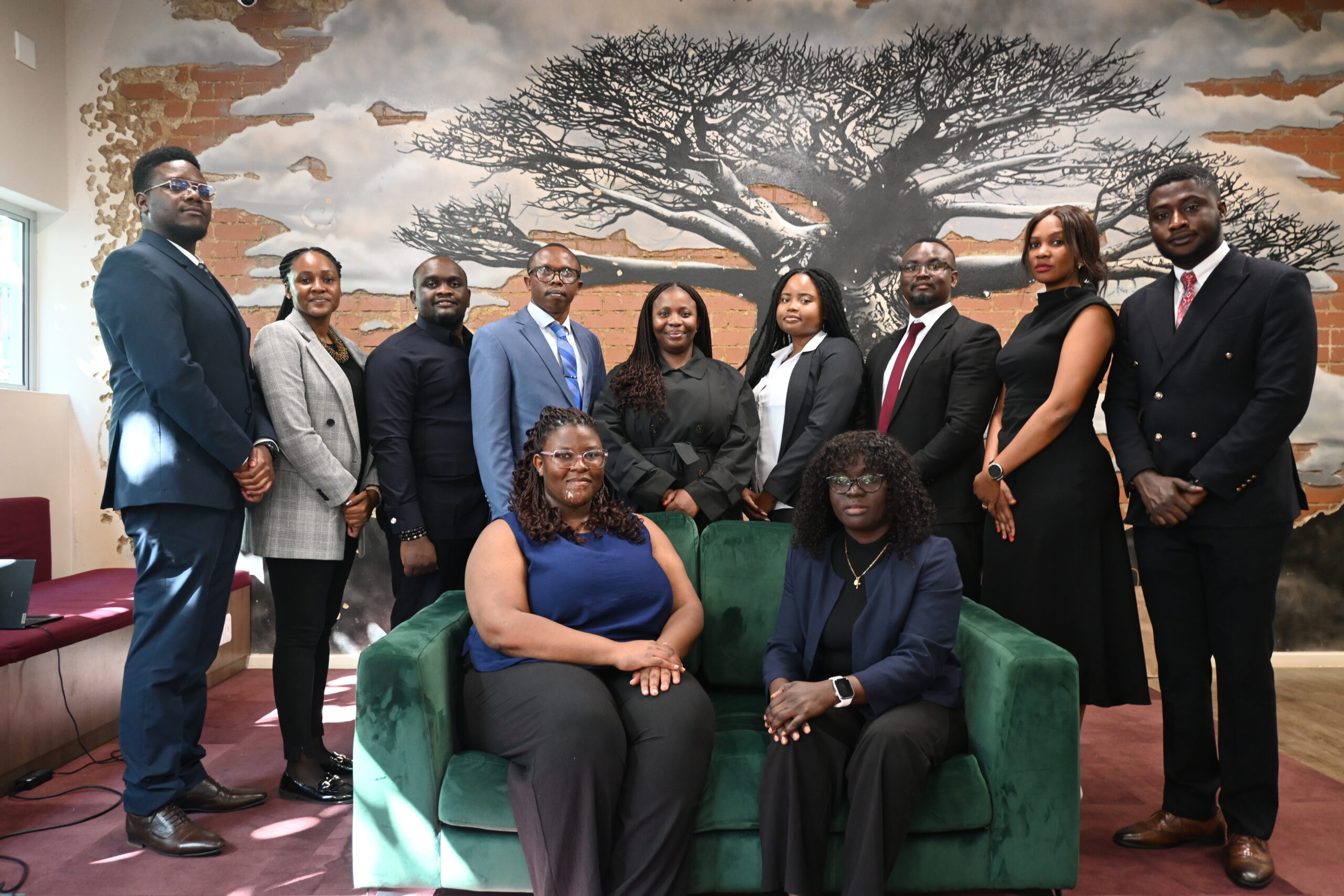 A group of eleven professionally dressed people pose indoors, with nine standing behind two seated on a green couch. Behind them is a large mural of a tree on a brick wall.
