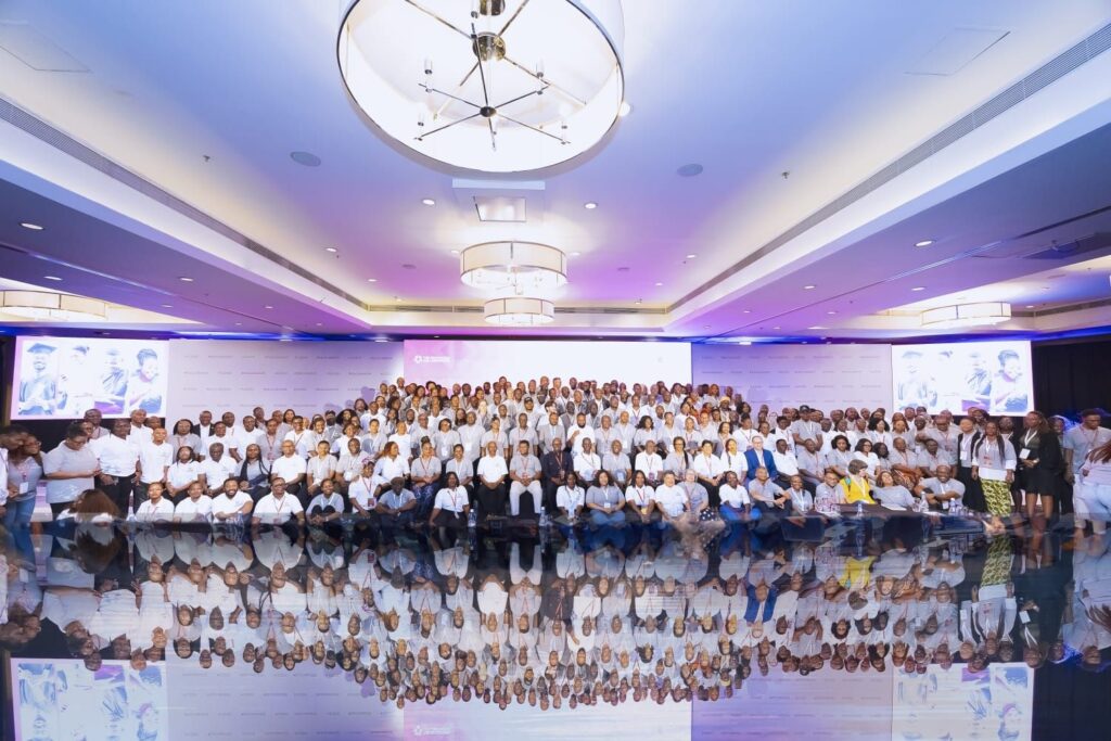 A large group of people from the Education Collaborative pose together in a brightly lit conference room with white walls and ceiling lights, all wearing white shirts, reflected on a shiny floor. A branded backdrop is behind them.