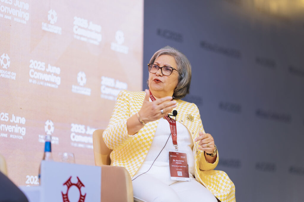 A woman in a yellow and white checkered blazer speaks on stage at an Education Collaborative conference, gesturing with her hand while holding a microphone. She wears glasses and a name badge, with â€œ2025 June Conveningâ€ displayed behind her.