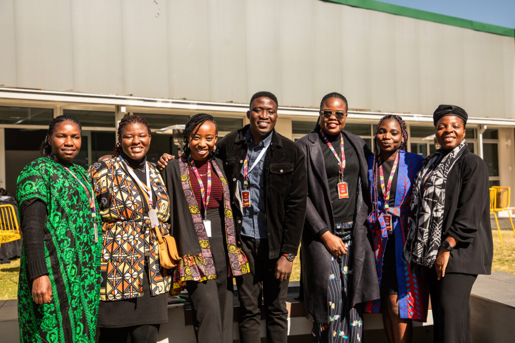 A group of seven people stand outdoors, smiling and posing for a photo. They are dressed in colorful, patterned clothing and wearing lanyards. The background features a modern building and yellow chairs.