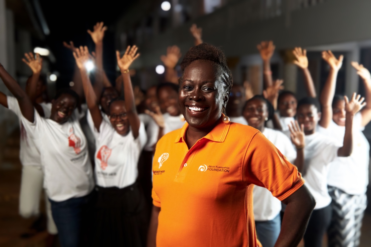 A smiling woman in an orange shirt stands in front of a group of people in white shirts, all raising their arms and cheering, outdoors at night. The group appears joyful and energetic.