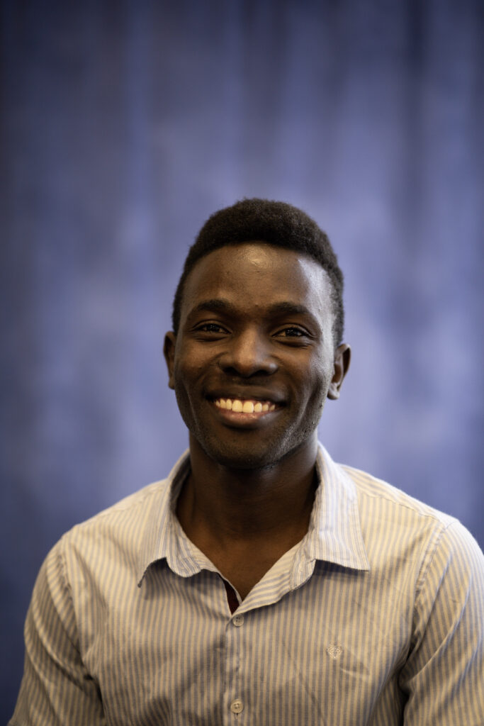 A man wearing a light striped shirt smiles at the camera with a blurred dark blue background behind him.