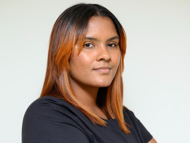 A woman with straight, shoulder-length auburn hair and a nose ring stands against a plain light background, wearing a black shirt and looking confidently at the camera.