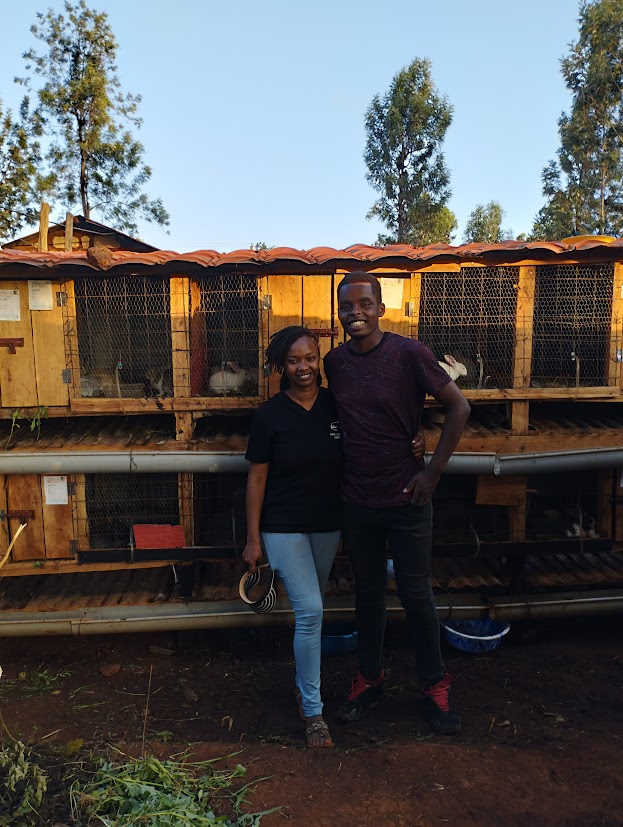 Two people smiling and standing arm-in-arm in front of wooden animal cages outdoors, with trees and blue sky in the background. The woman holds a hat and both look happy.