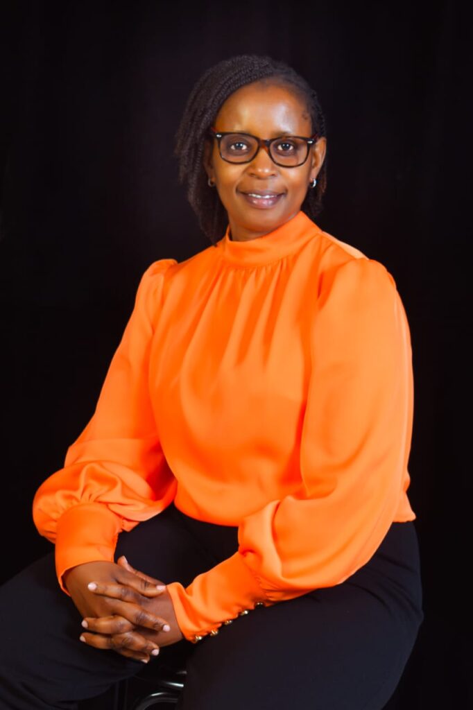 A woman with short braided hair, wearing glasses, an orange long-sleeve blouse, and black pants sits on a stool with her hands folded, against a plain black background.
