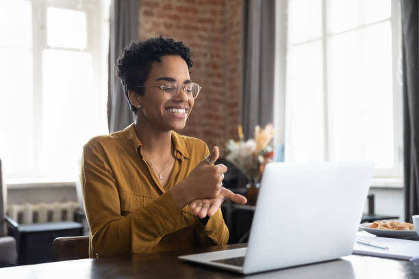 A smiling person wearing glasses and a brown shirt sits at a desk, using sign language while looking at a laptop in a well-lit room with large windows.